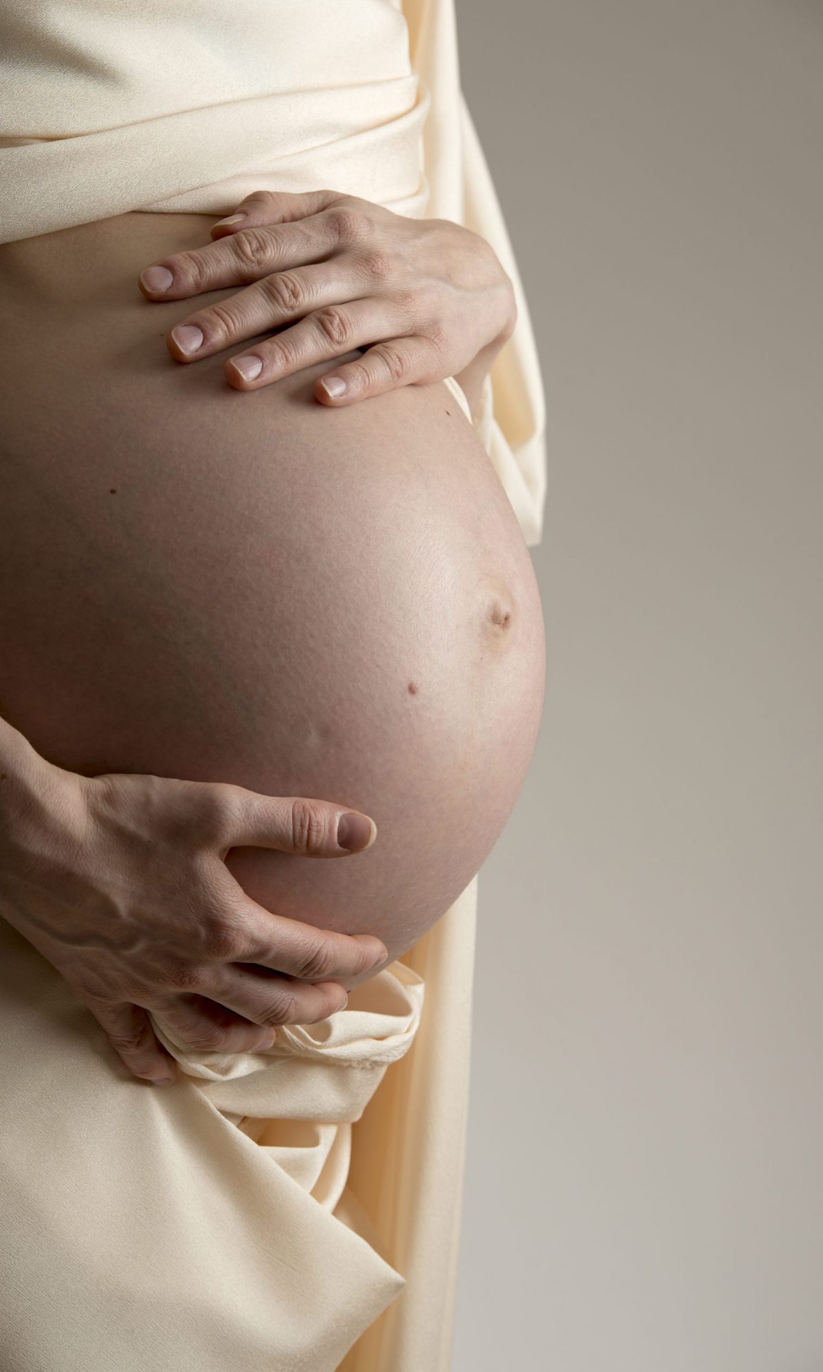 Women in dressing gown opens it to hold her pregnant abdomen with hand and fingers, shaded cream background.